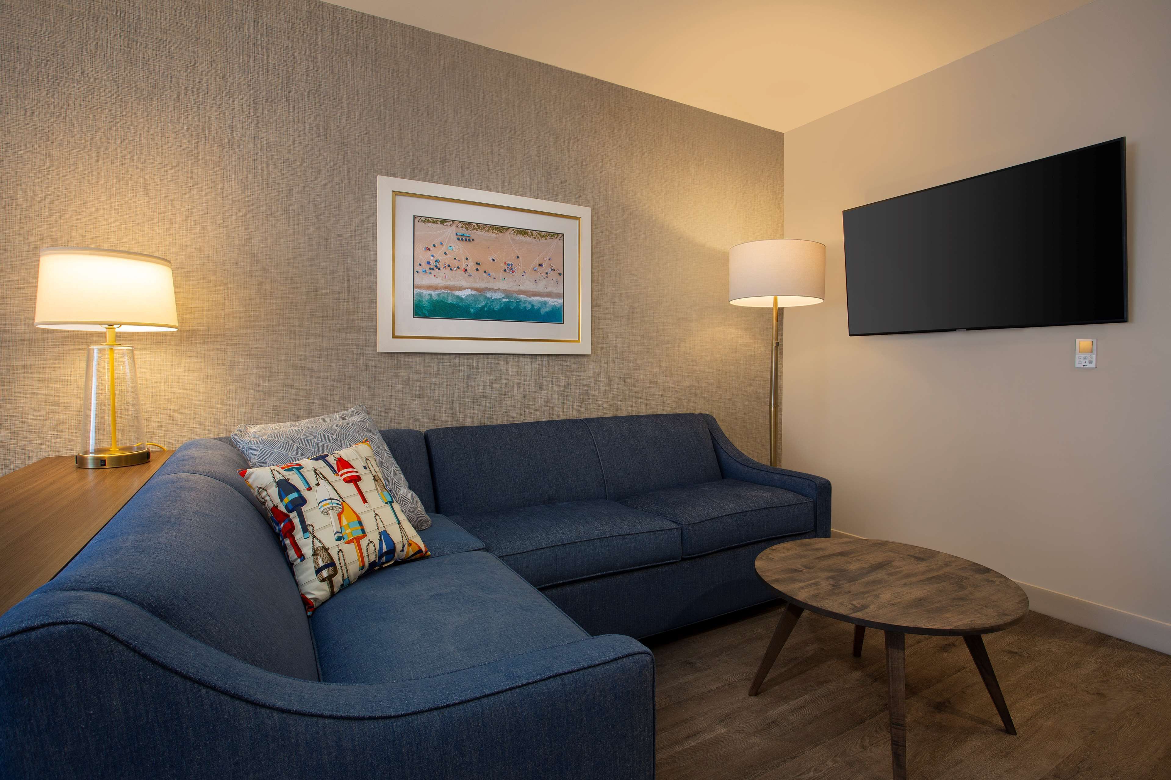 A modern living room with a blue sofa, a small round table, two floor lamps, and a flat-screen TV mounted on the wall.