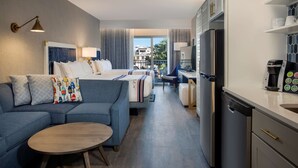 Une chambre d’hôtel moderne équipée d’un canapé, d’une petite table, d’un lit avec une tête de lit, d’un coin cuisine avec un réfrigérateur et une machine à café, et offrant une vue sur l’extérieur.