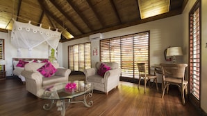 Un salón espacioso con suelo de madera, una mesa de centro de cristal, dos sofás beige con cojines rosa, una silla de mimbre y ventanas grandes con persianas.