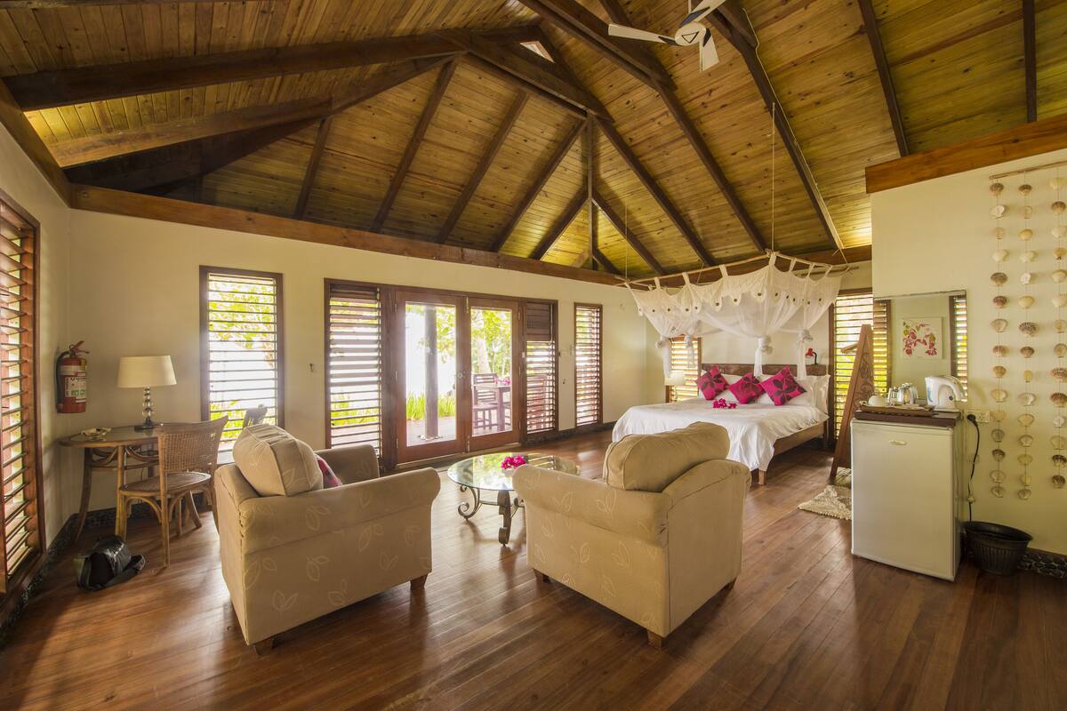 A spacious room with wooden floors, a bed with a canopy, two sofas, a small table, and a refrigerator.