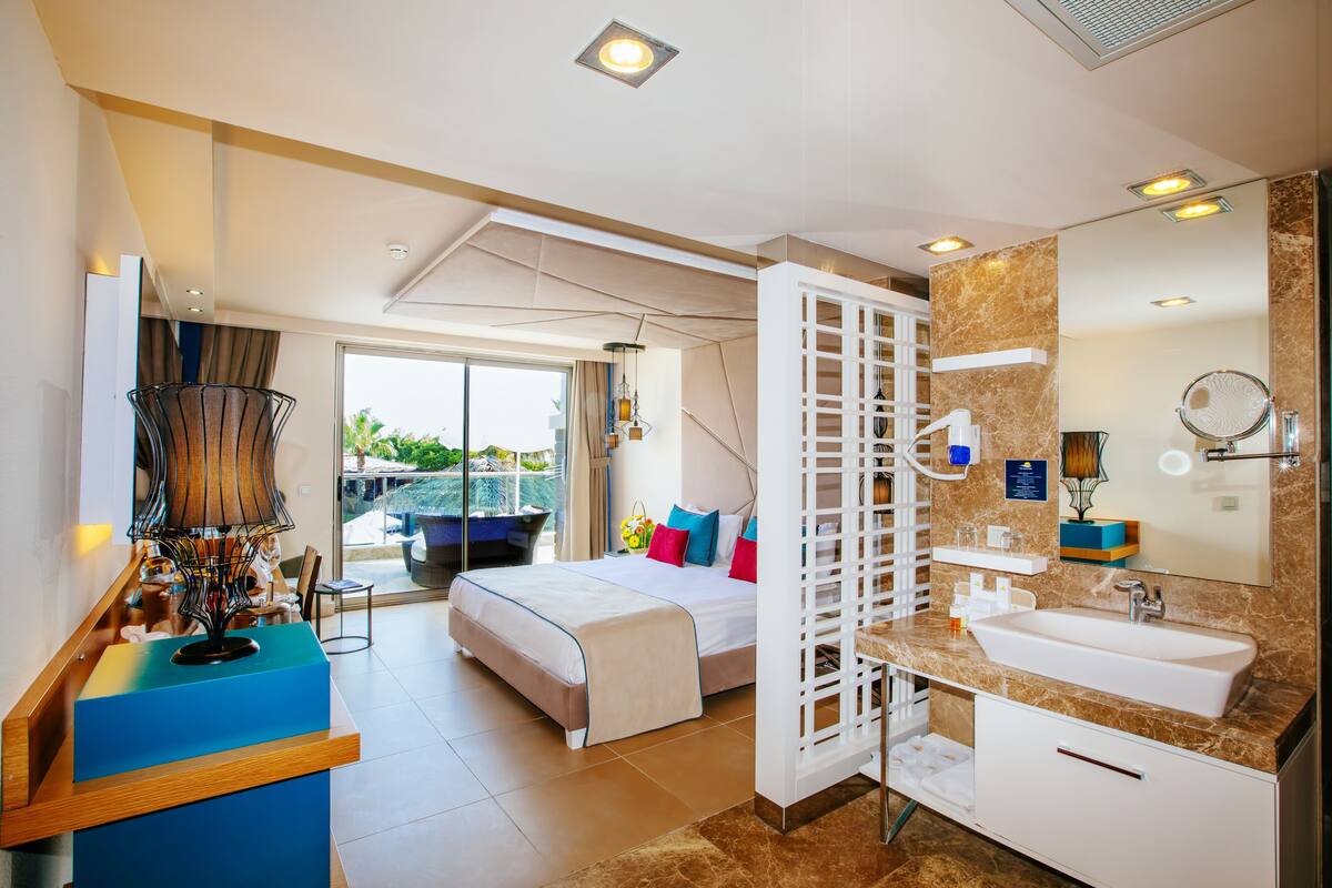 Ein modernes Hotelzimmer mit einem großen Bett, einem Badezimmer mit Waschbecken und Spiegel sowie einem Balkon mit Blick.