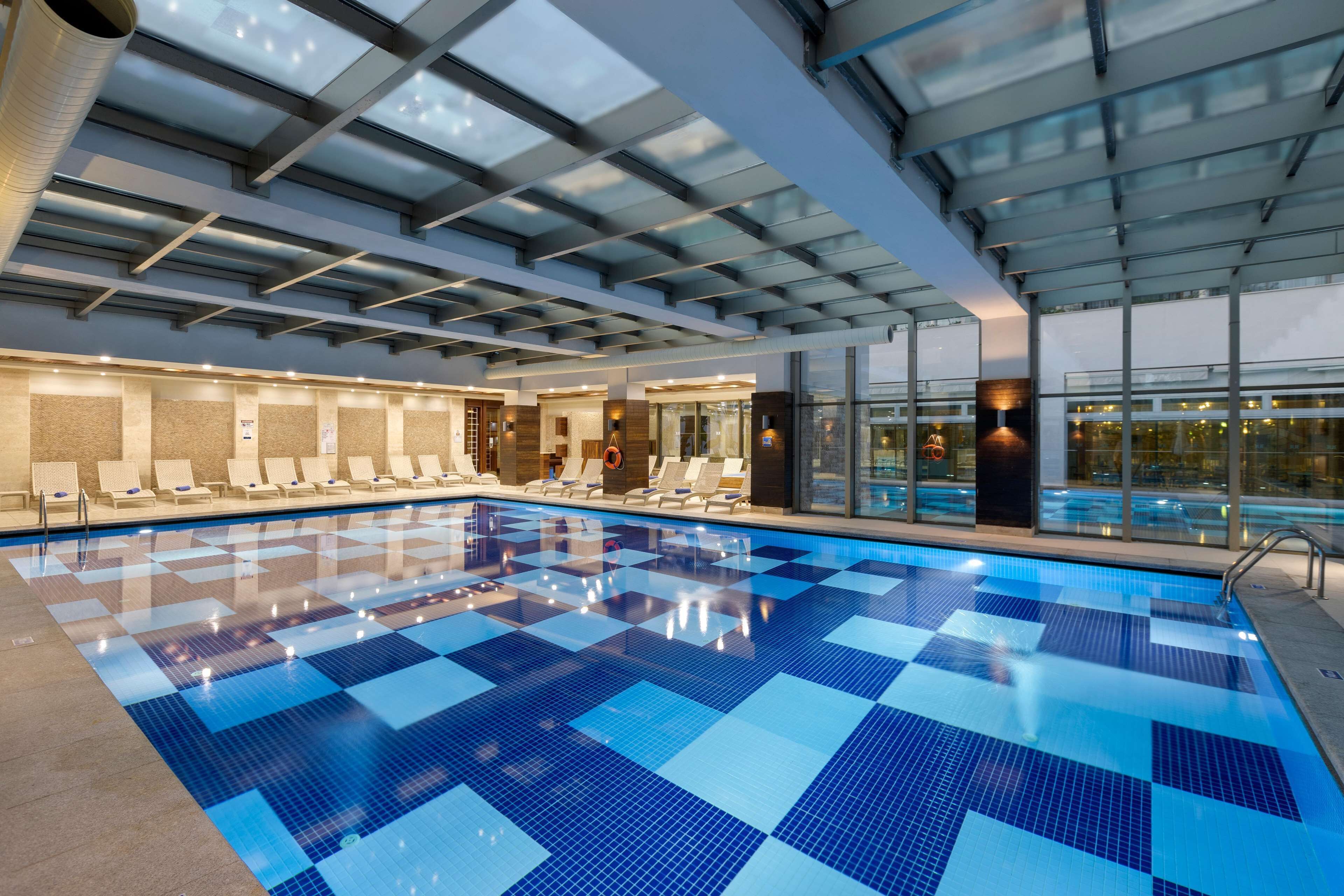 A large indoor swimming pool with a checkered blue tile pattern, sunroofed ceiling, and lounge chairs along the poolside.