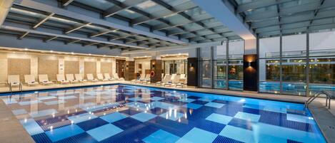 A large indoor swimming pool with a checkered blue tile pattern, sunroofed ceiling, and lounge chairs along the poolside.