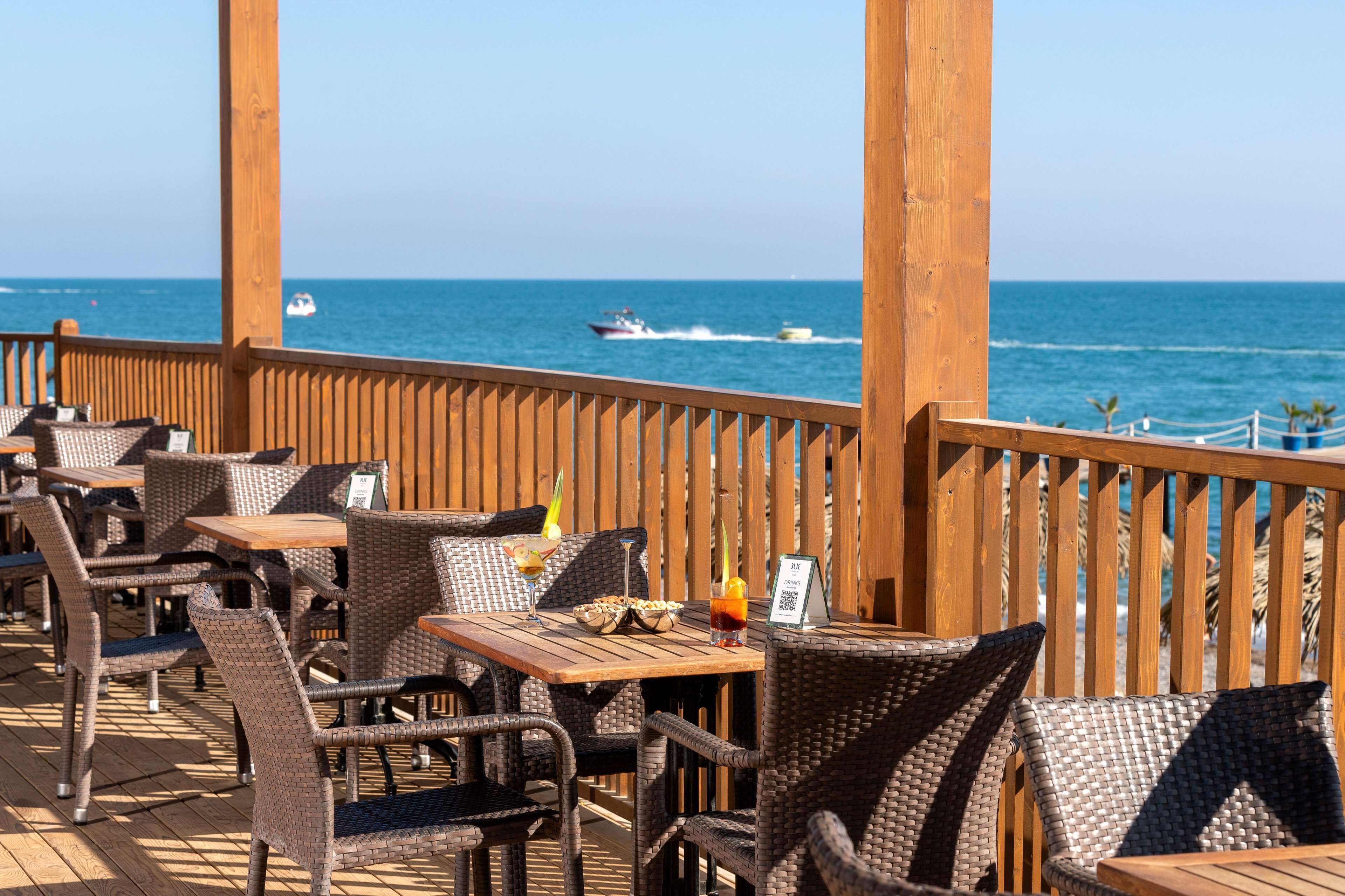 Un espace de restauration en plein air, doté de mobilier en osier et de tables en bois, offrant une vue sur la mer, avec un bateau visible au loin.