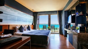 A hotel room with a large bed, a desk, a TV, and a balcony with a view of the ocean.