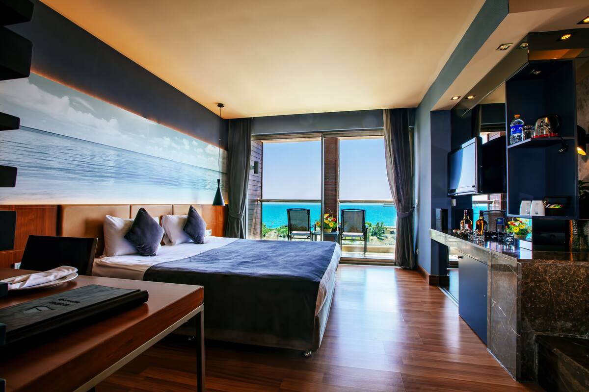 Ein Hotelzimmer mit einem großen Bett, einem Schreibtisch, einem Fernseher und einem Balkon mit Meerblick.