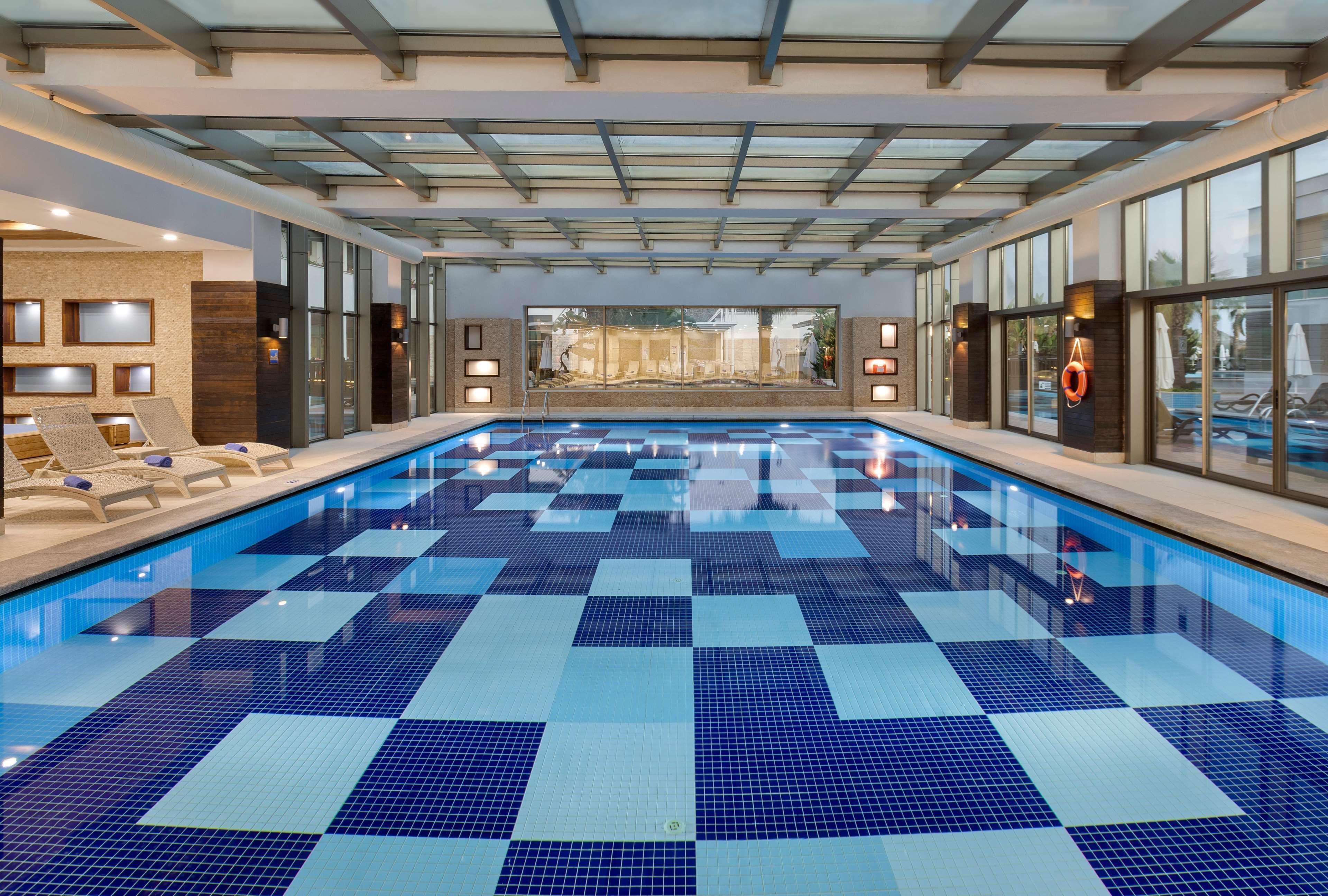 A large indoor swimming pool with checkered blue and white tiles, surrounded by lounge chairs and large windows.
