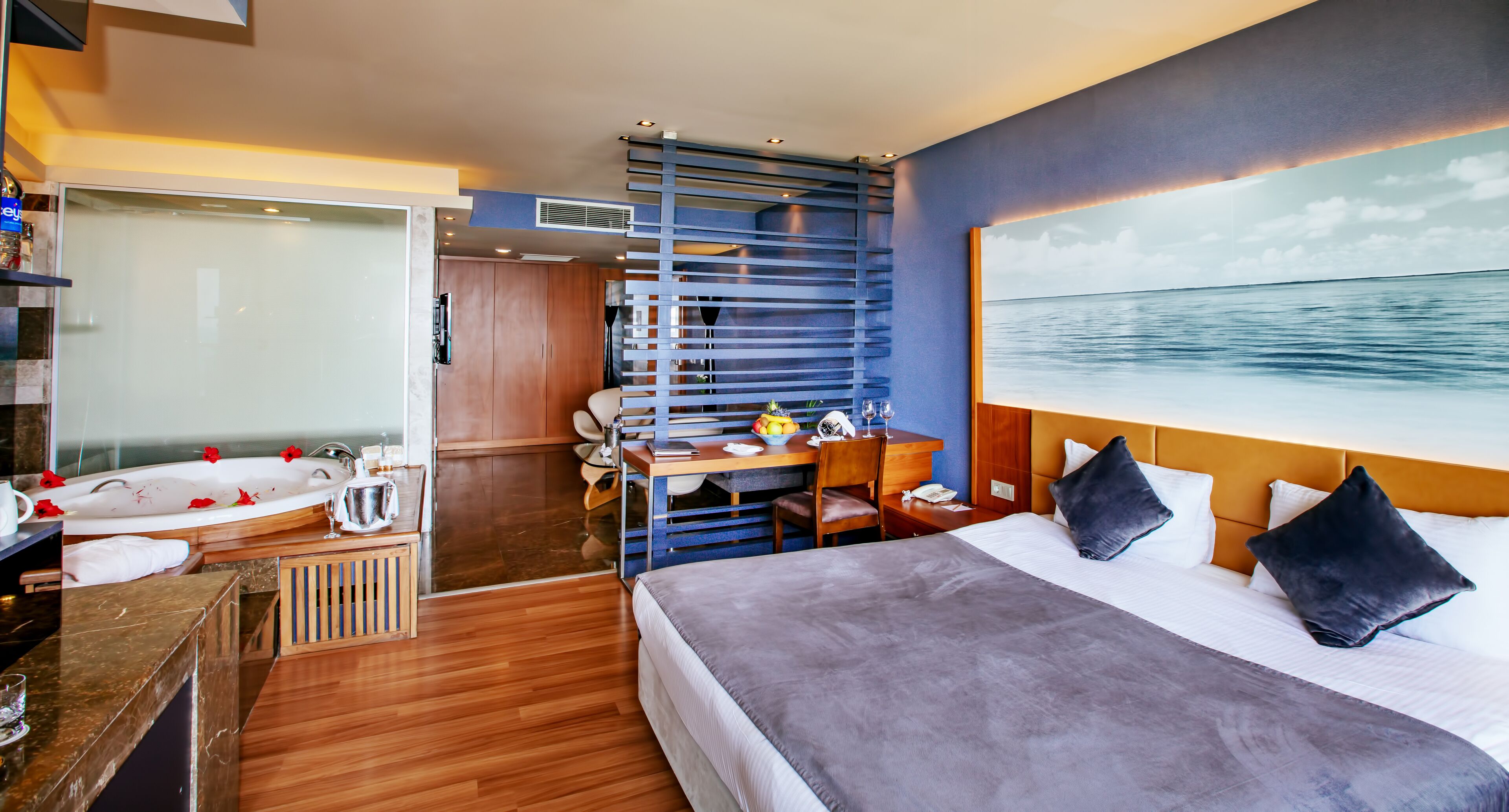 A modern hotel room with a large bed, a desk with a chair, a small seating area, a bathtub, and a large window with a view of the sea.
