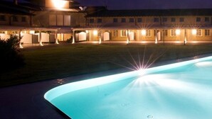 Une piscine illuminée la nuit, dont les lumières se reflètent sur l’eau, entourée d’un bâtiment doté de nombreuses fenêtres et d’une façade bien éclairée.