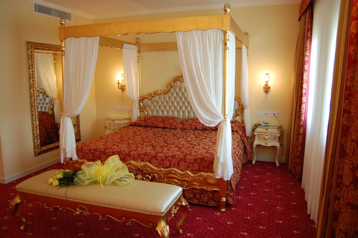 A four-poster bed with white drapes, a red patterned bedspread, and a gold-framed headboard. There is a bedside table with a lamp, a mirror with a gold frame, and a red carpeted floor.