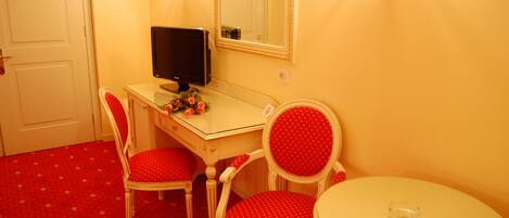 A room with a desk, two red upholstered chairs, a round table, a mirror, and a television.