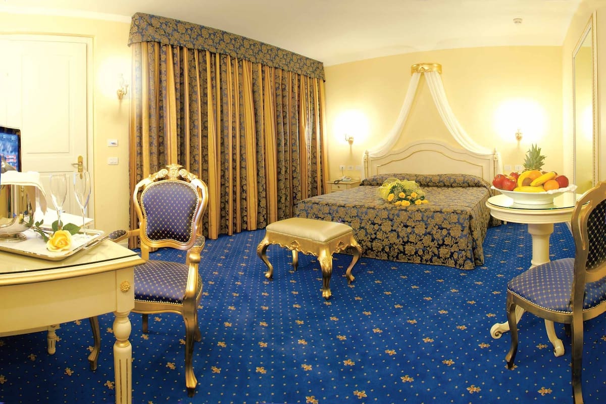 A hotel room with a large bed, a desk with a computer, a chair, a small table with fruit, and a canopy bed.