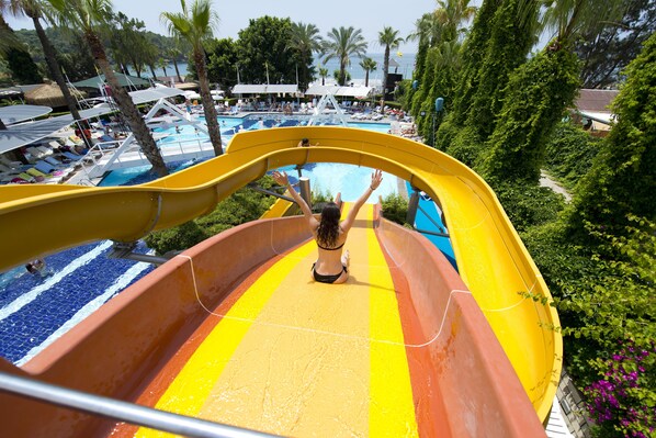 Un toboggan aquatique avec une personne en haut, entouré d’une zone de piscine avec des chaises longues et des parasols.