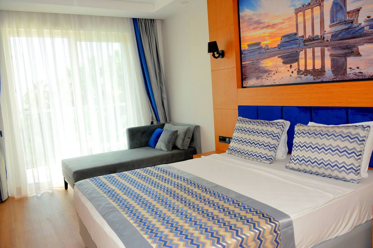 Een hotelkamer met een groot bed, een kleinere slaapbank, een schilderij van een historisch gebouw en een raam met dunne gordijnen.