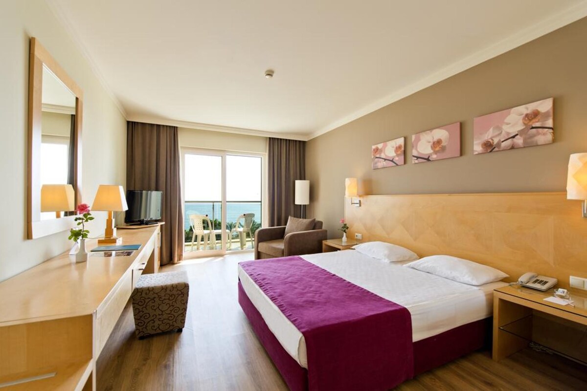 A hotel room with a large bed, a desk with a lamp, a chair, a TV, and artwork on the wall.