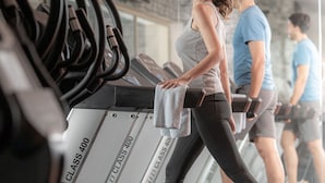 Uma academia com esteiras, uma pessoa usando uma esteira e outra pessoa ao fundo.