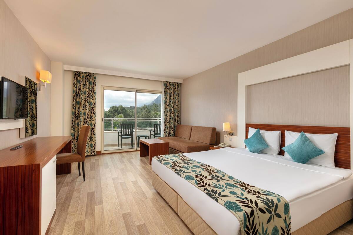 A hotel room with a large bed, a desk, a chair, a sofa, and a balcony with a view of greenery.