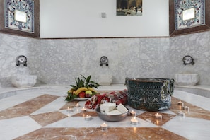 Un baño con piso de mármol, un frutero con fruta, velas y una pileta decorativa.
