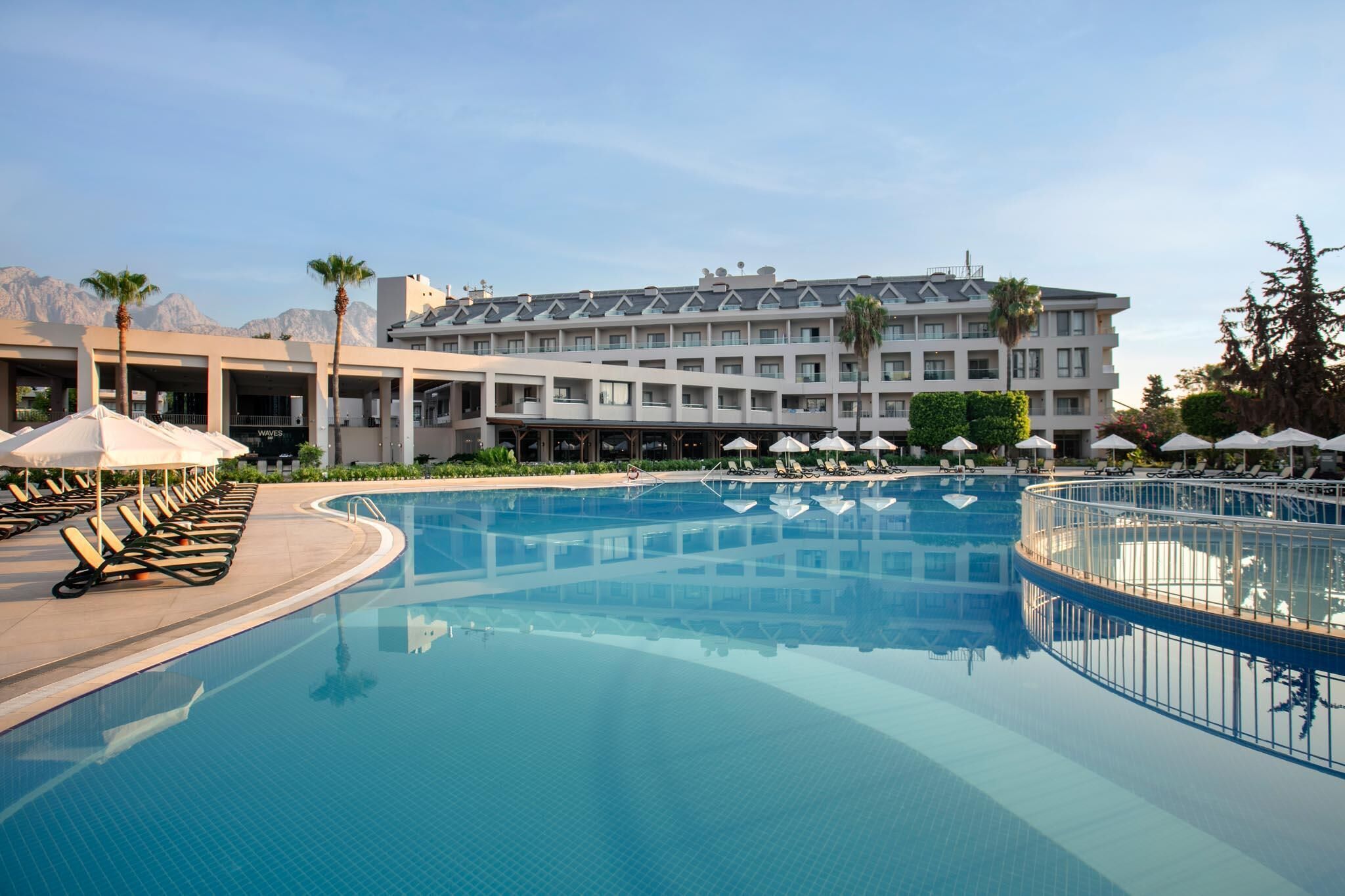 Una piscina con sillones de descanso y sombrillas, un edificio de hotel con varios pisos y balcones, y palmeras.