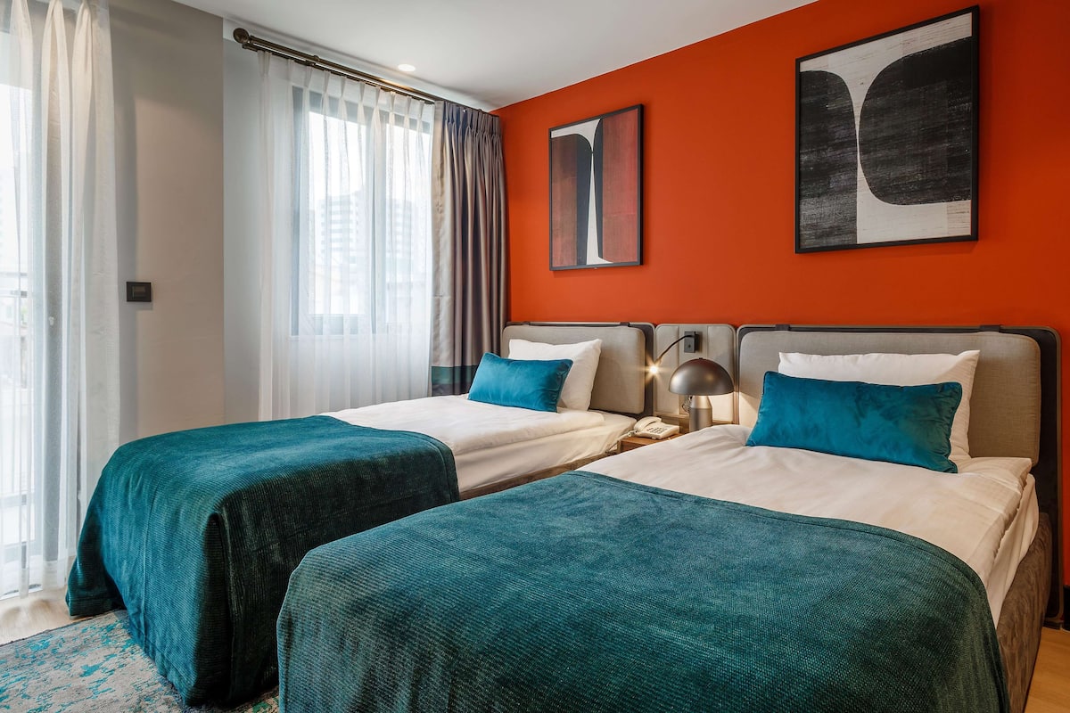 Quarto de hotel com duas camas, uma janela grande com cortinas e uma parede de destaque laranja vibrante.