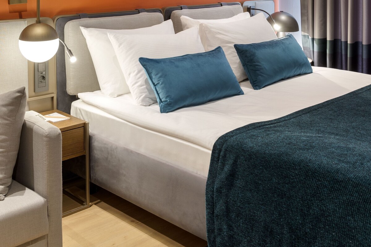 A modern hotel room with a large bed, a bedside table, a lamp, and artwork on the wall.