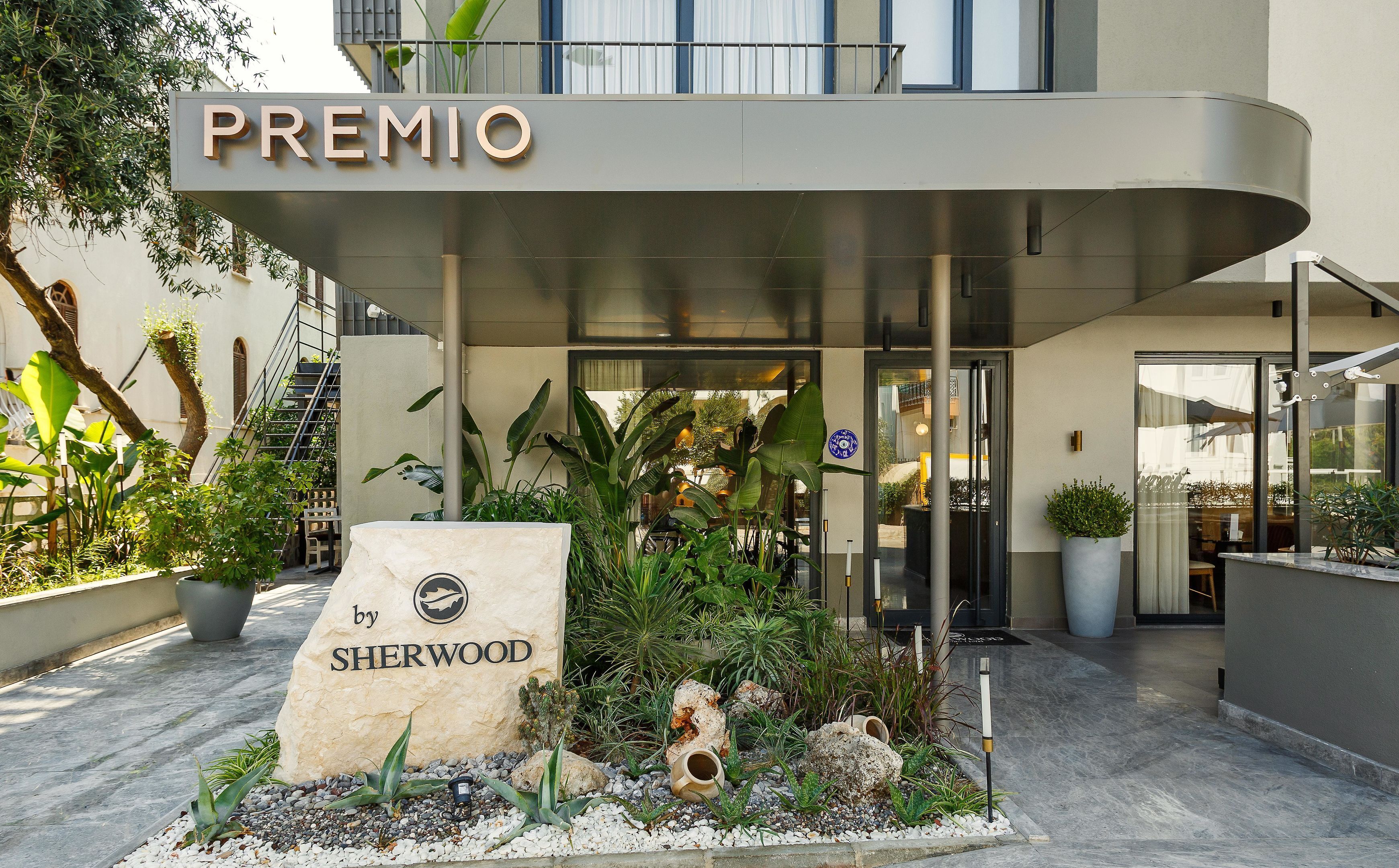 L’entrée d’un hôtel, avec une enseigne indiquant « PREMIO by SHERWOOD », et un espace paysager agrémenté de plantes et de rochers.