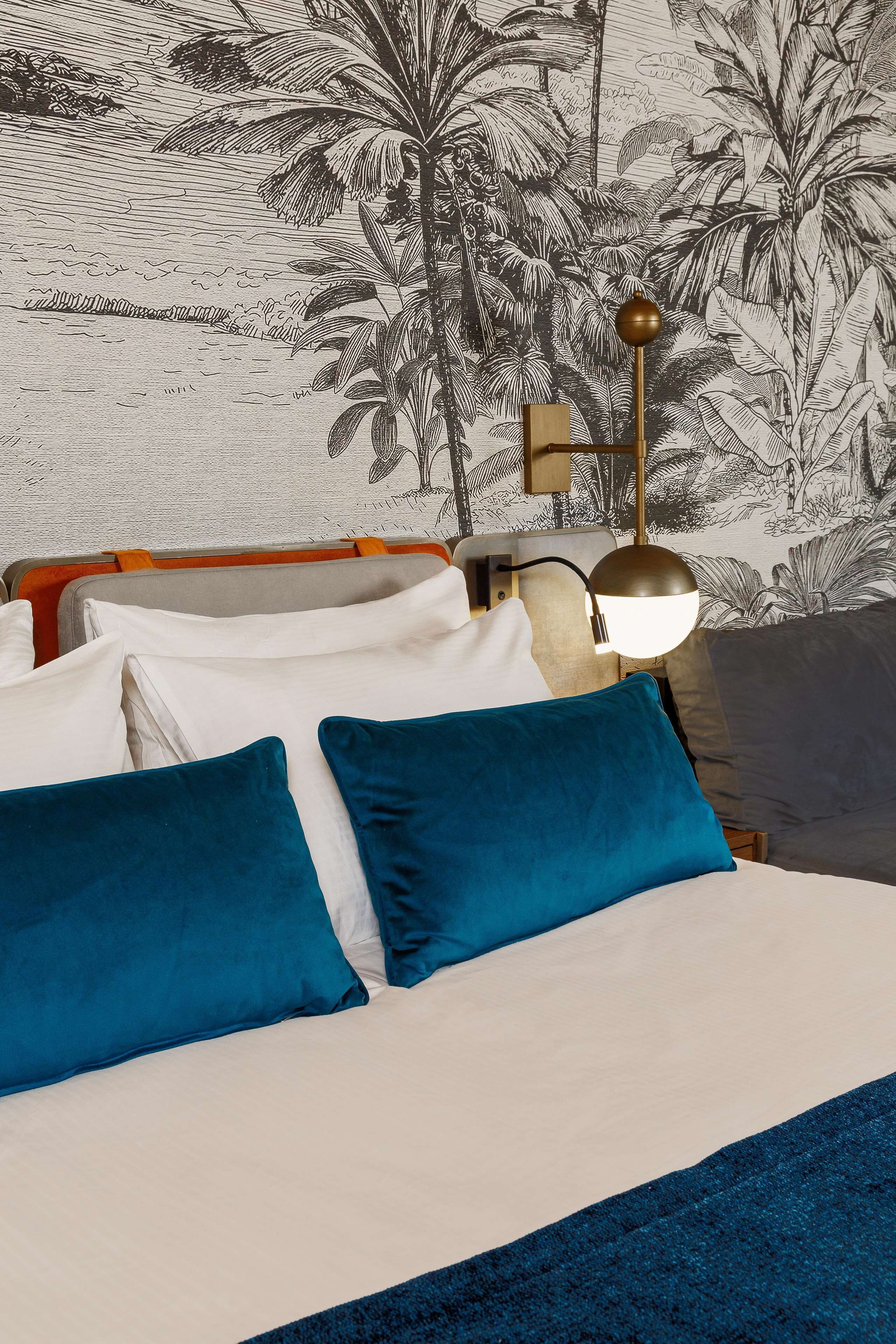 A bed with blue and white pillows, a nightstand with a lamp, and a tropical wallpaper.