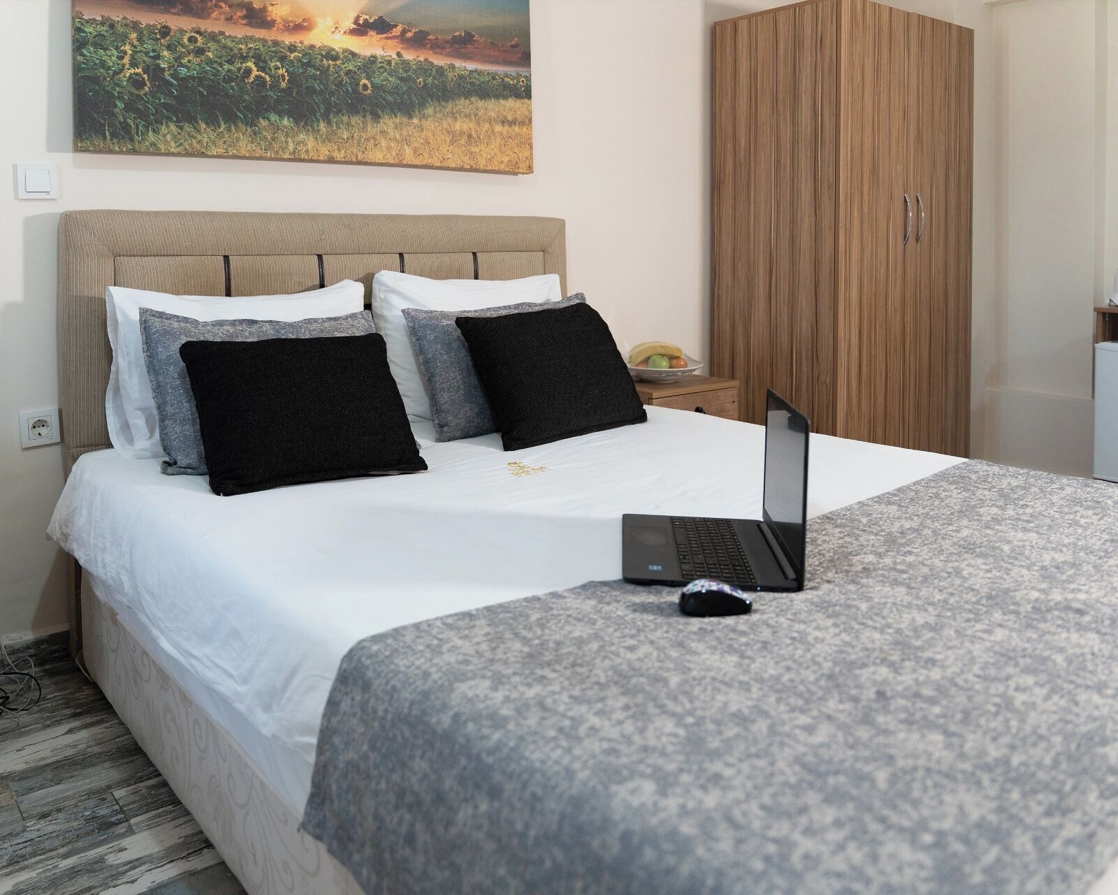 Una cama bien hecha con un ordenador portátil encima, un armario de madera y un cuadro de un campo en la pared.