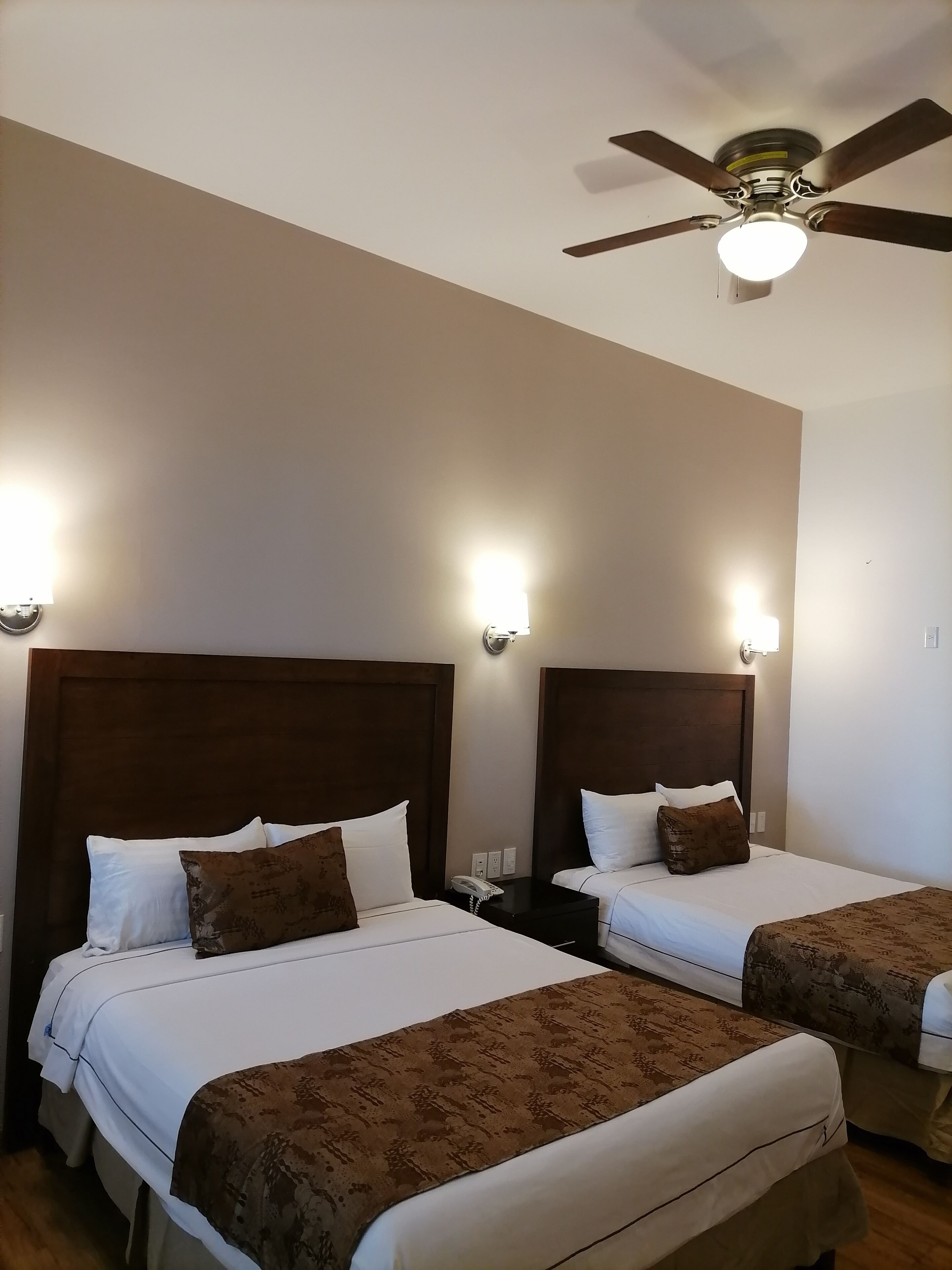 Habitación de hotel con dos camas, cada una con cabecero de madera, ropa de cama blanca y una manta marrón con estampado. Hay lámparas de pared y un ventilador de techo.