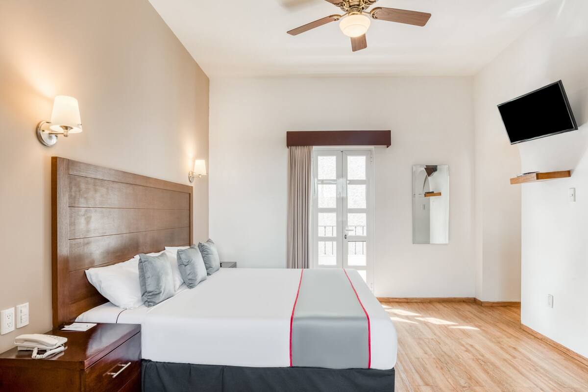 Una habitación de hotel con cabecera de madera, cama con edredón blanco y gris, ventilador de techo, televisor montado en la pared y ventana con cortinas.