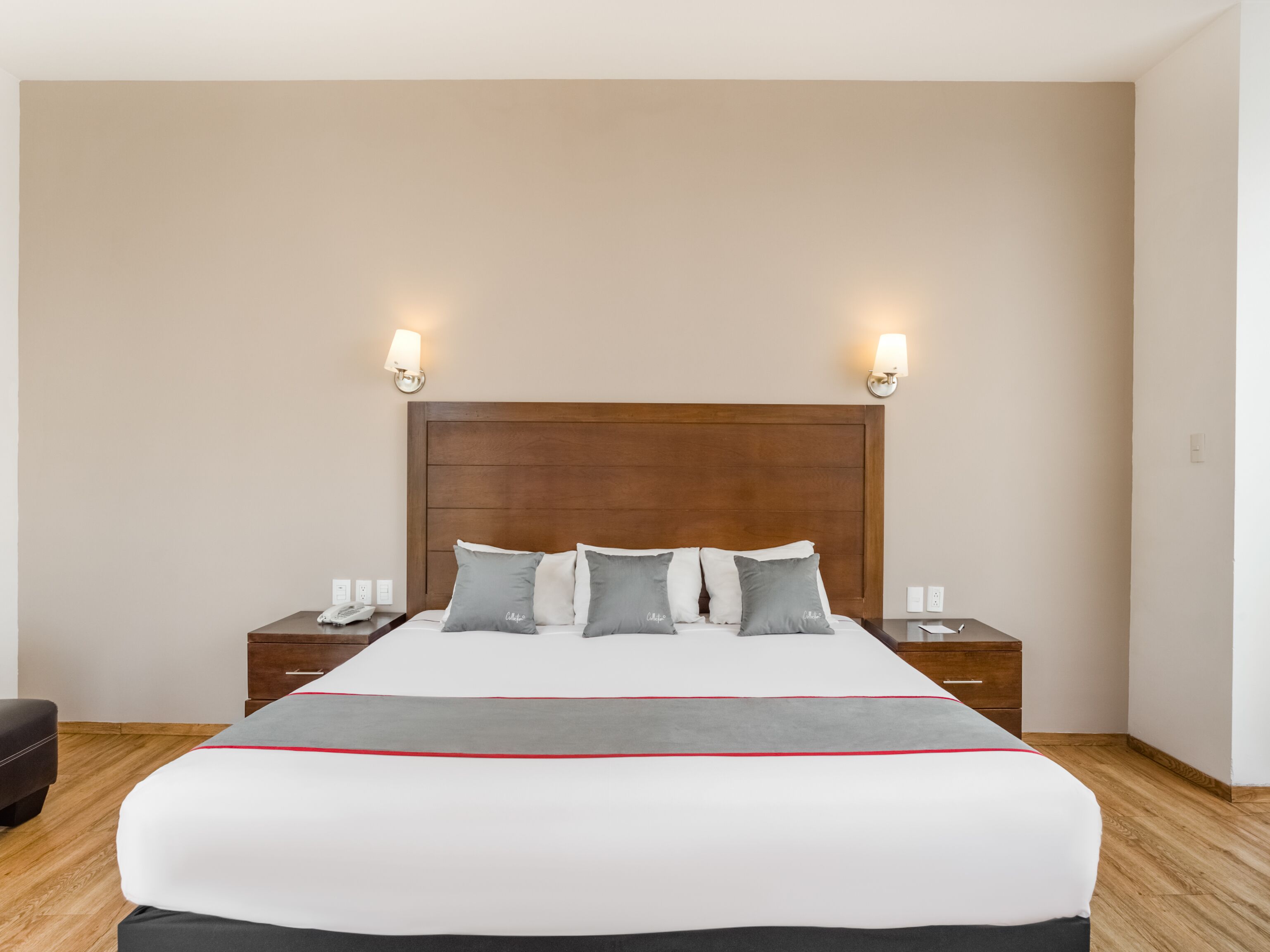A hotel room with a large bed, wooden headboard, two bedside tables with lamps, and a small nightstand.