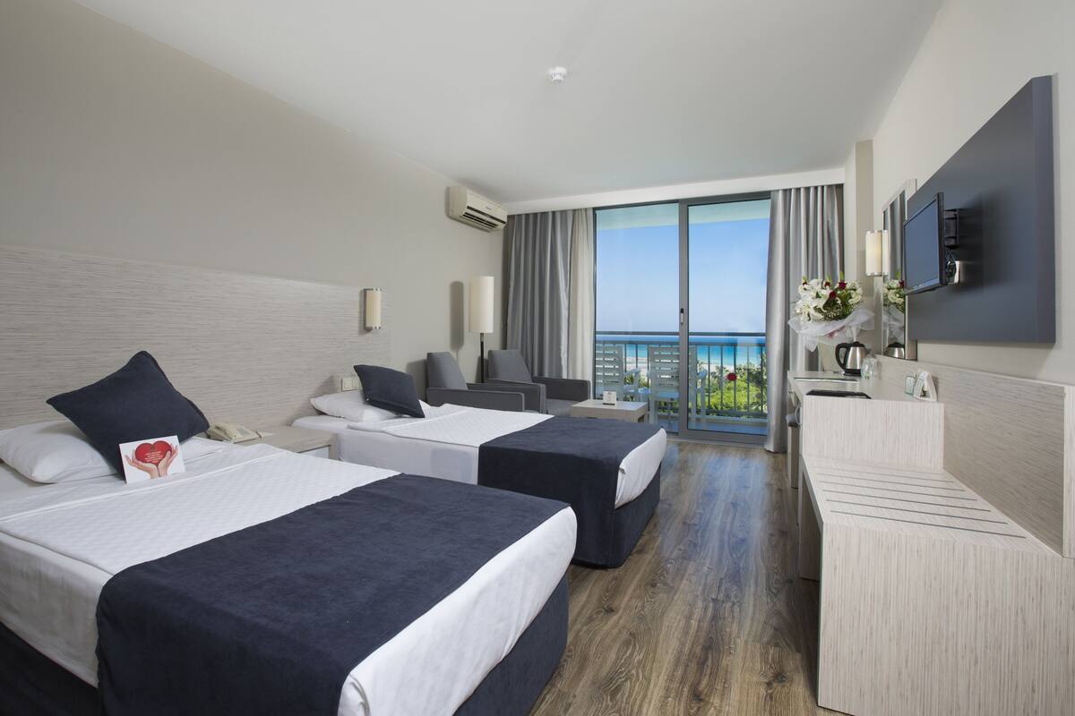 A hotel room with two beds, a desk, a chair, a TV, and a balcony with a view of the sea.