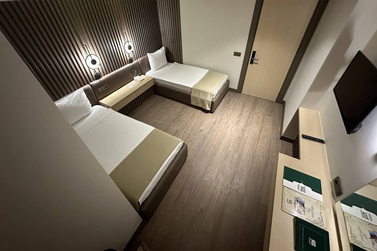 A compact hotel room with two single beds, a small desk, a wall-mounted TV, and a wooden floor.