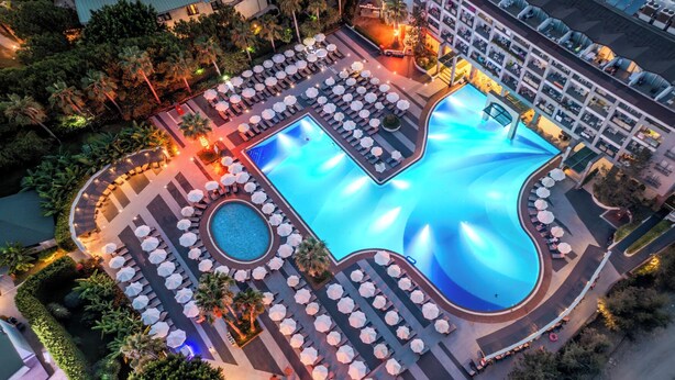 Ein großer Außenpool mit mehreren Liegestühlen und Sonnenschirmen, umgeben von Grünflächen und einem mehrstöckigen Hotelgebäude.