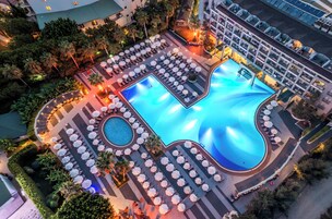 Una amplia piscina al aire libre con múltiples tumbonas y sombrillas, rodeada de follaje cuidado y un edificio de hotel de varios pisos.