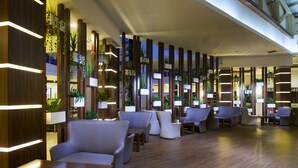 Eine moderne Lobby mit Holzwandverkleidung, vertikalen Lichtleisten und Blumentöpfen. Es gibt mehrere Sitzbereiche mit Tischen und Stühlen.