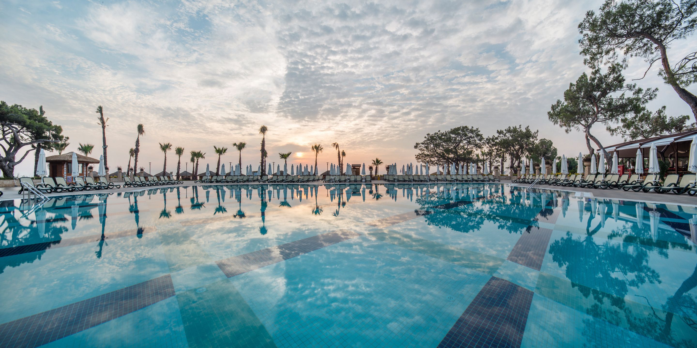 A swimming pool with lounge chairs and umbrellas, surrounded by trees and a sunset sky.