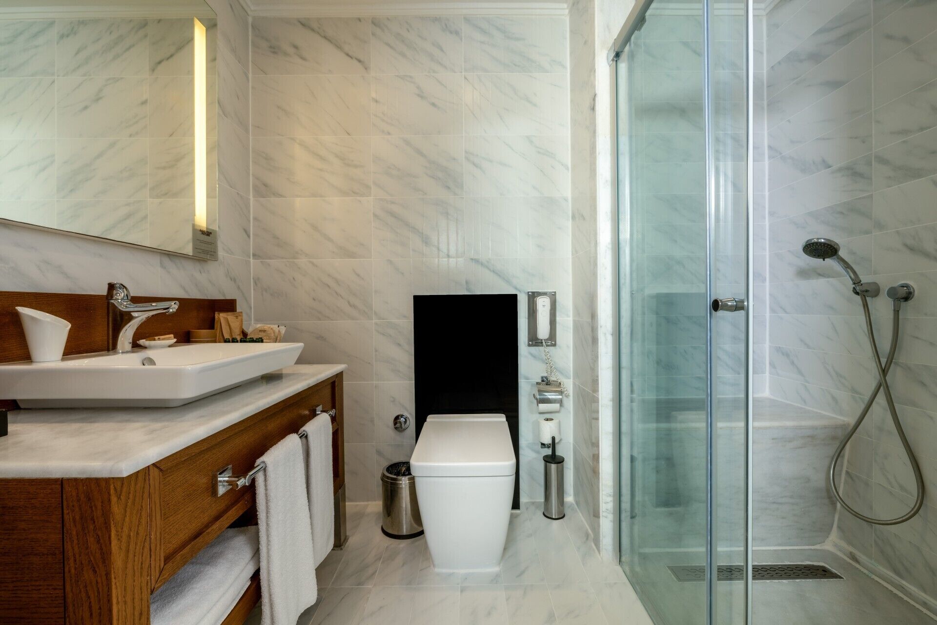 A modern bathroom with a marble floor, a wooden vanity, a glass shower enclosure, and a toilet.