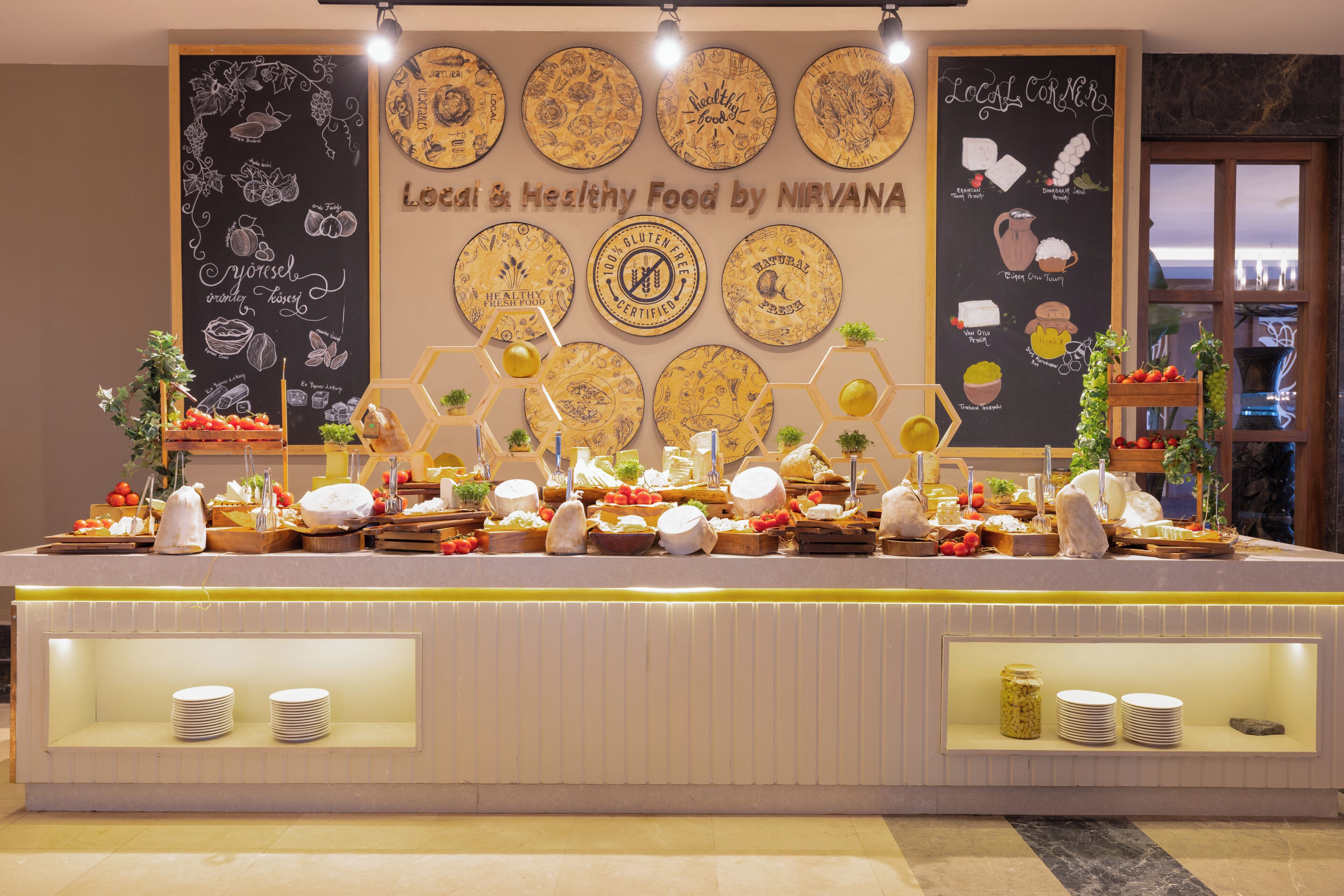 Un mostrador de buffet con diferentes alimentos, una pizarra con dibujos relacionados con la comida y un cartel que promociona comida local y saludable.