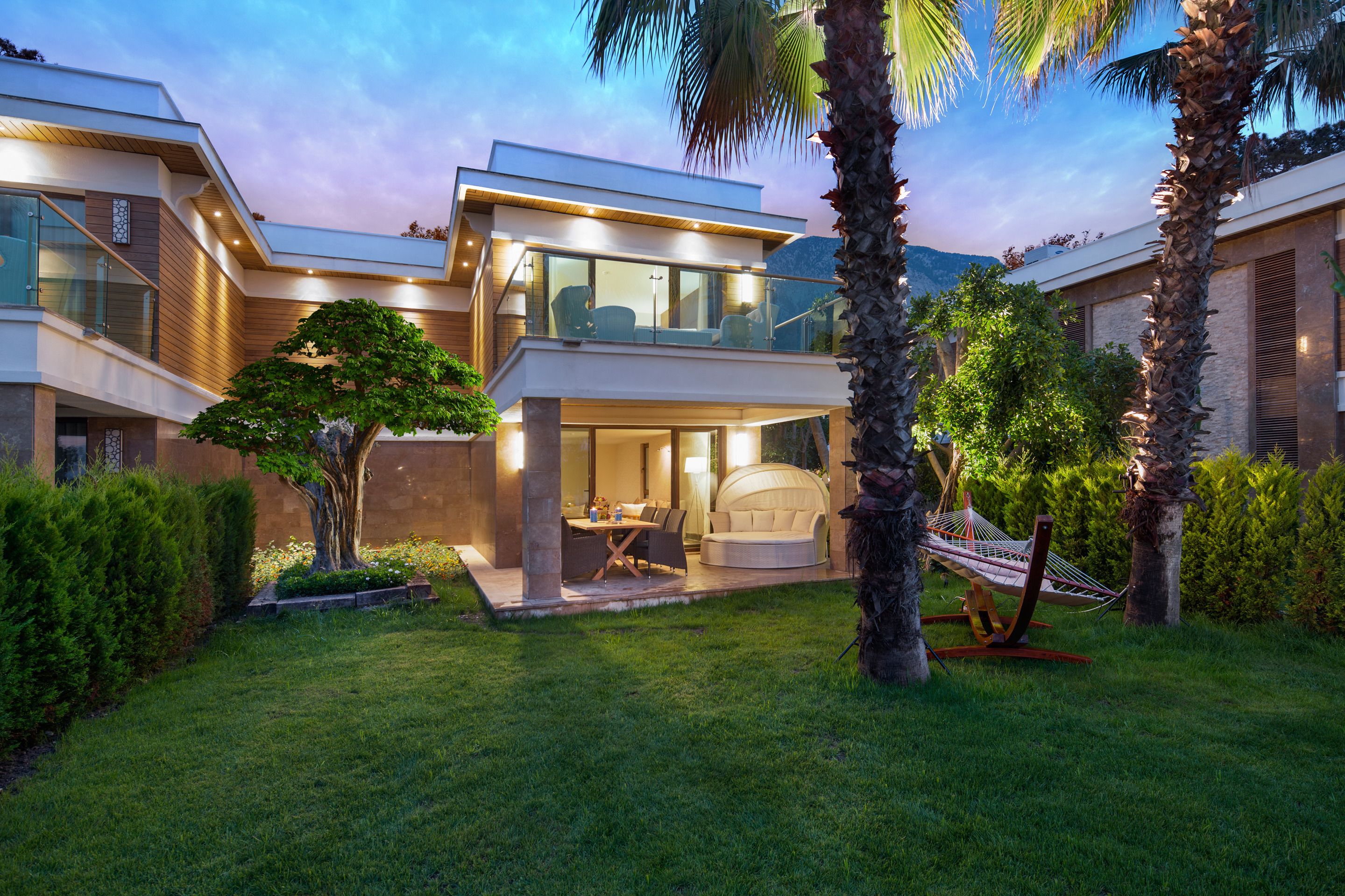 A modern two-story house with a balcony, a dining area, and a hammock in the garden.