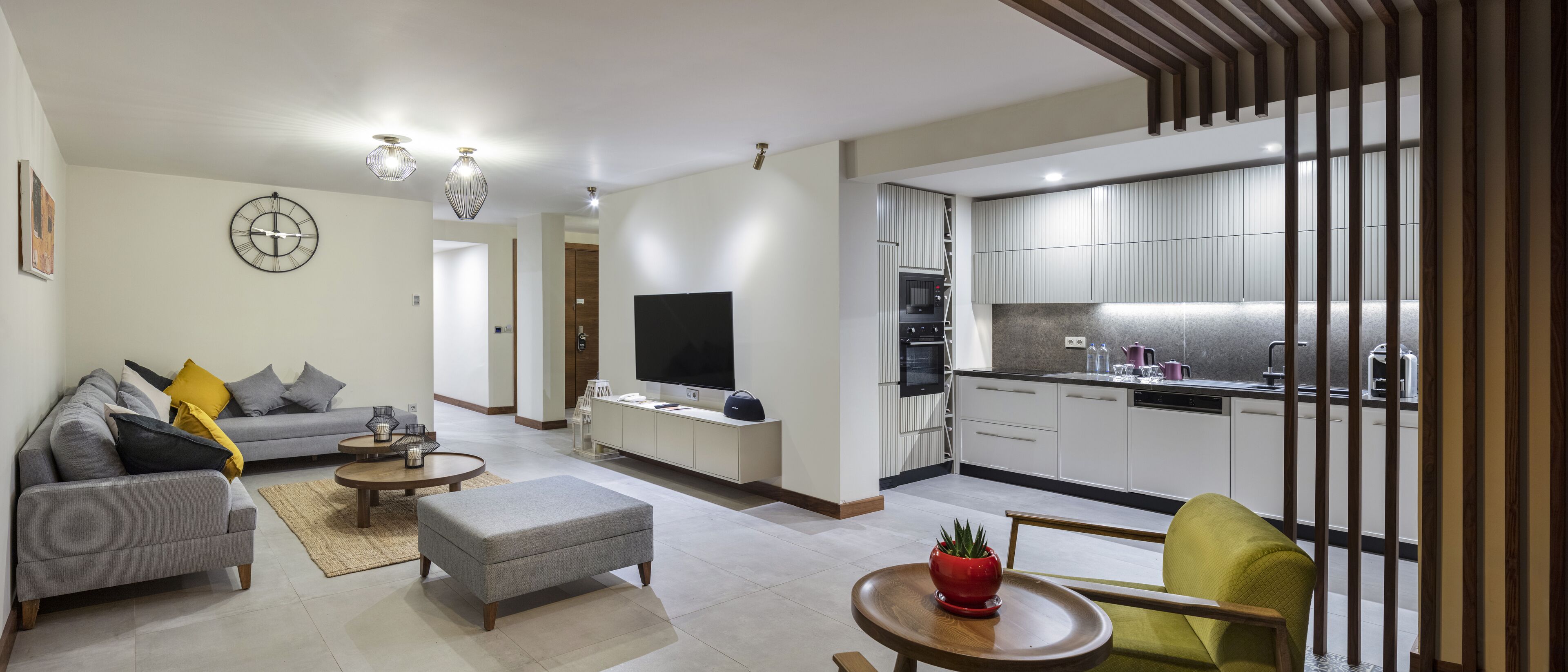 Een moderne woonkamer met een grijze bank, een poef en een flatscreen televisie. Een keuken met witte kasten en een eethoek met een houten tafel en stoelen is zichtbaar.