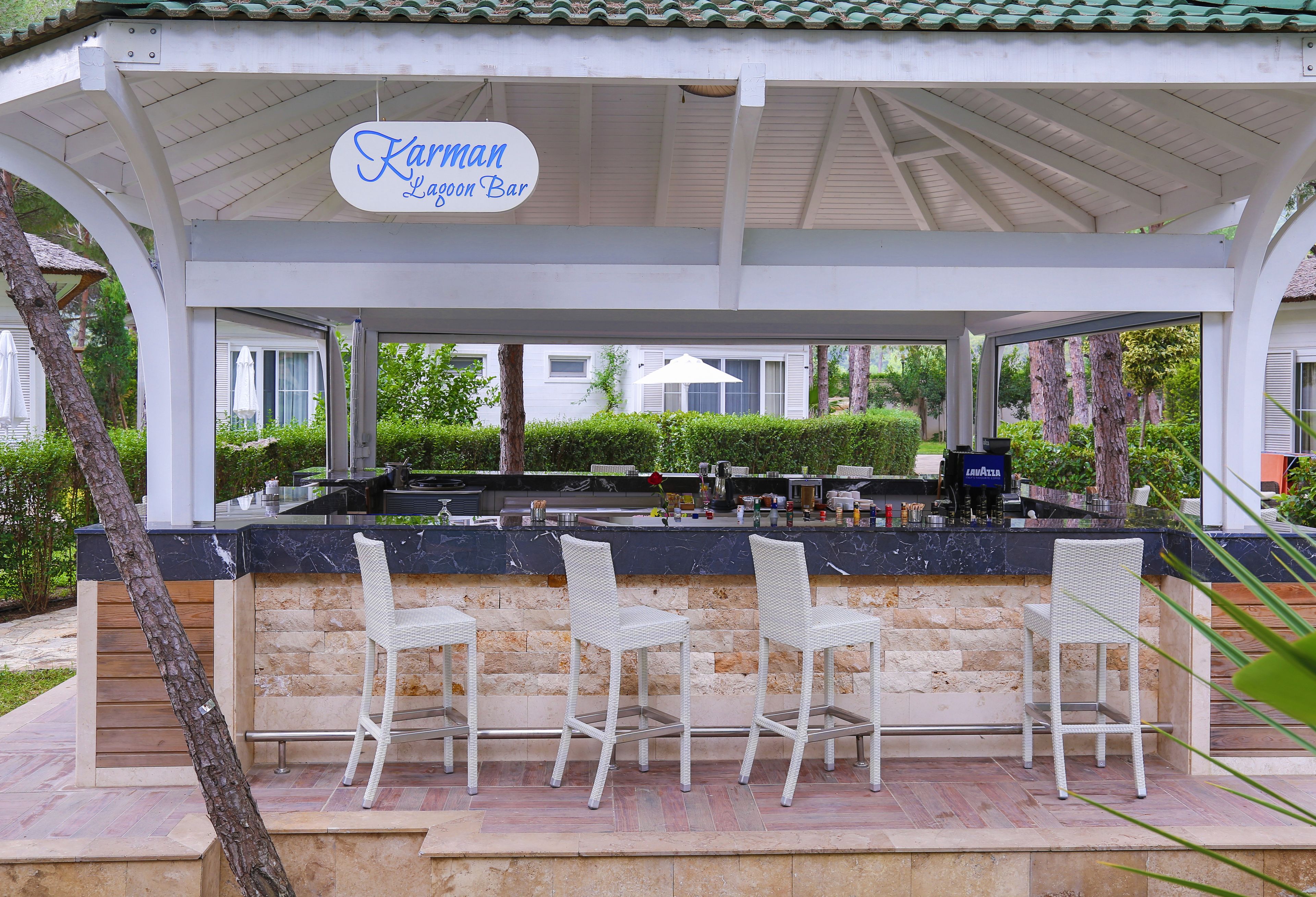 Zona de bar al aire libre cubierta con barra de piedra, sillas de mimbre blanco y un letrero que dice "Karman Lagoon Bar".