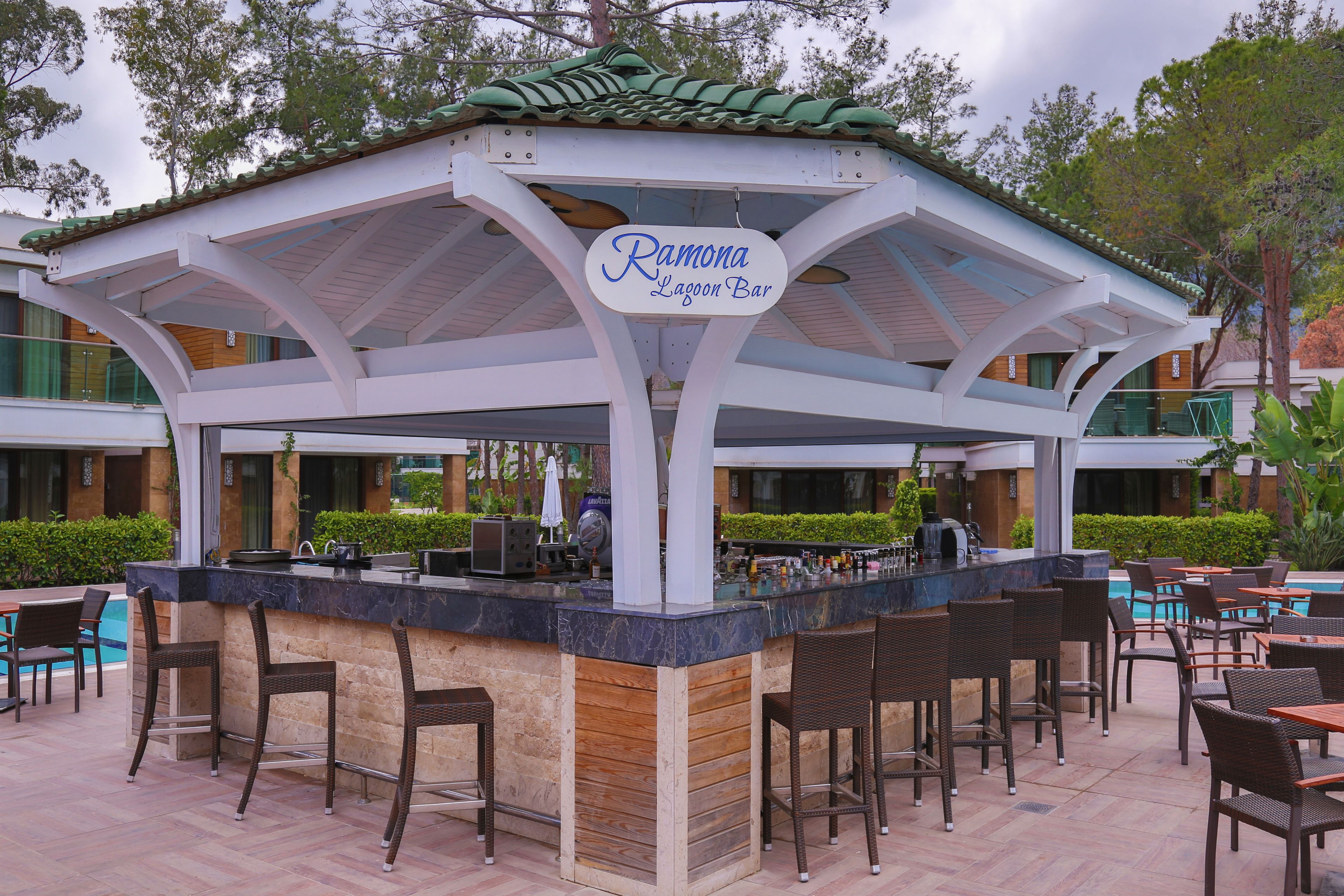 Uma área de bar ao ar livre coberta com uma placa que diz "Ramona Lagoon Bar", com balcão, banquetas e mesas para sentar.