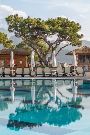 Ein Poolbereich mit Liegestühlen und Sonnenschirmen, ein Holzbau und ein großer Baum.