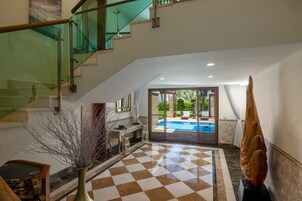 A spacious indoor area with a checkered floor, a staircase with glass railings, a pool visible through glass doors, and a modern sculpture.