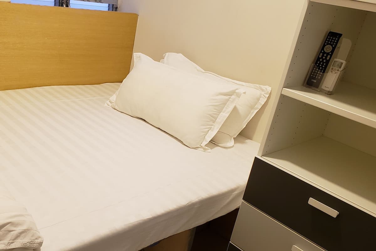 A neatly made bed with white linens and pillows, a wooden headboard, and a bedside table with a remote control and a charging cable.