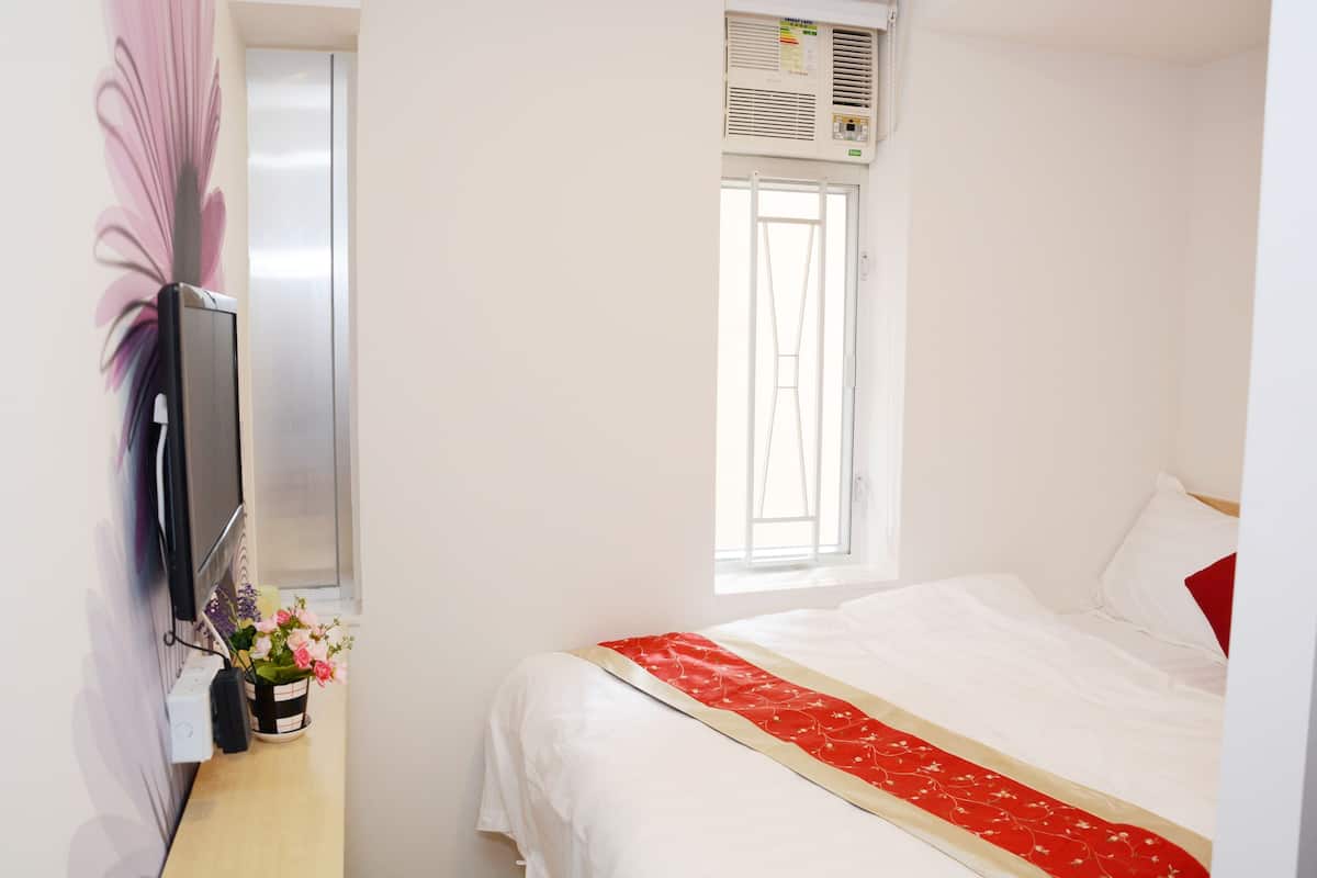 A hotel room with a bed, a TV, a window, and a wall-mounted air conditioner.