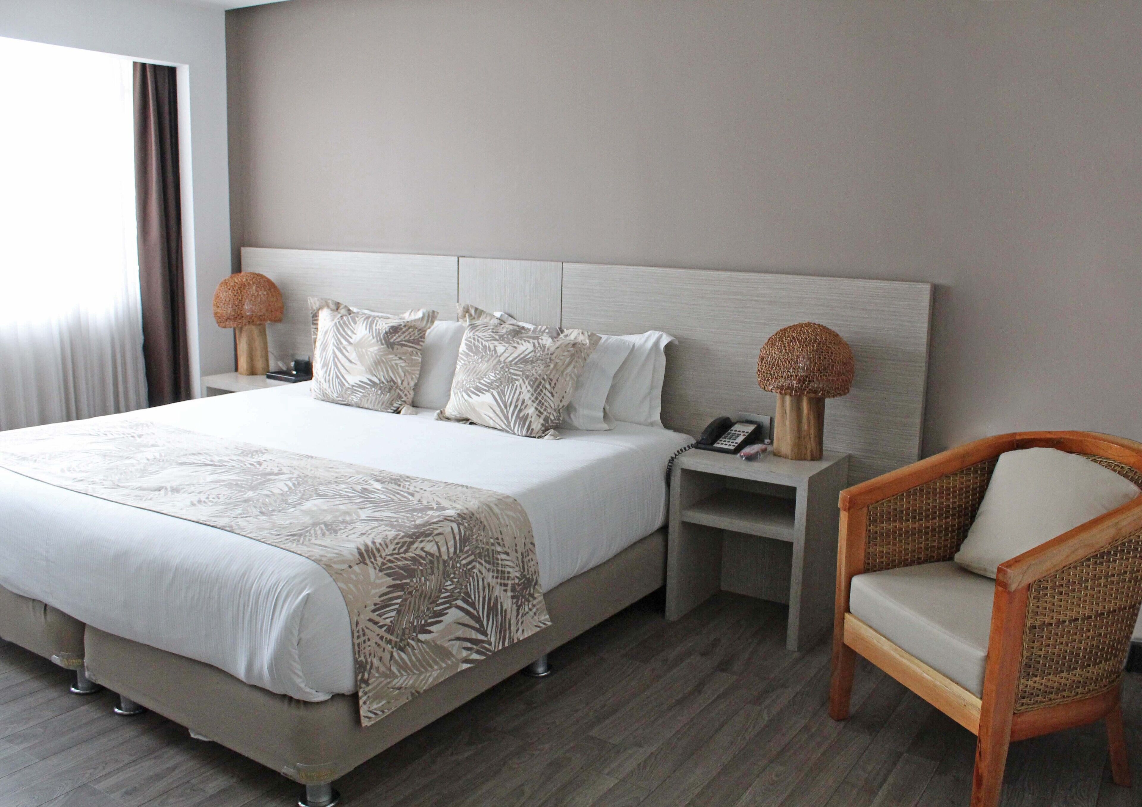 Quarto de hotel com uma cama grande, mesa de cabeceira com luminária e cadeira de vime.