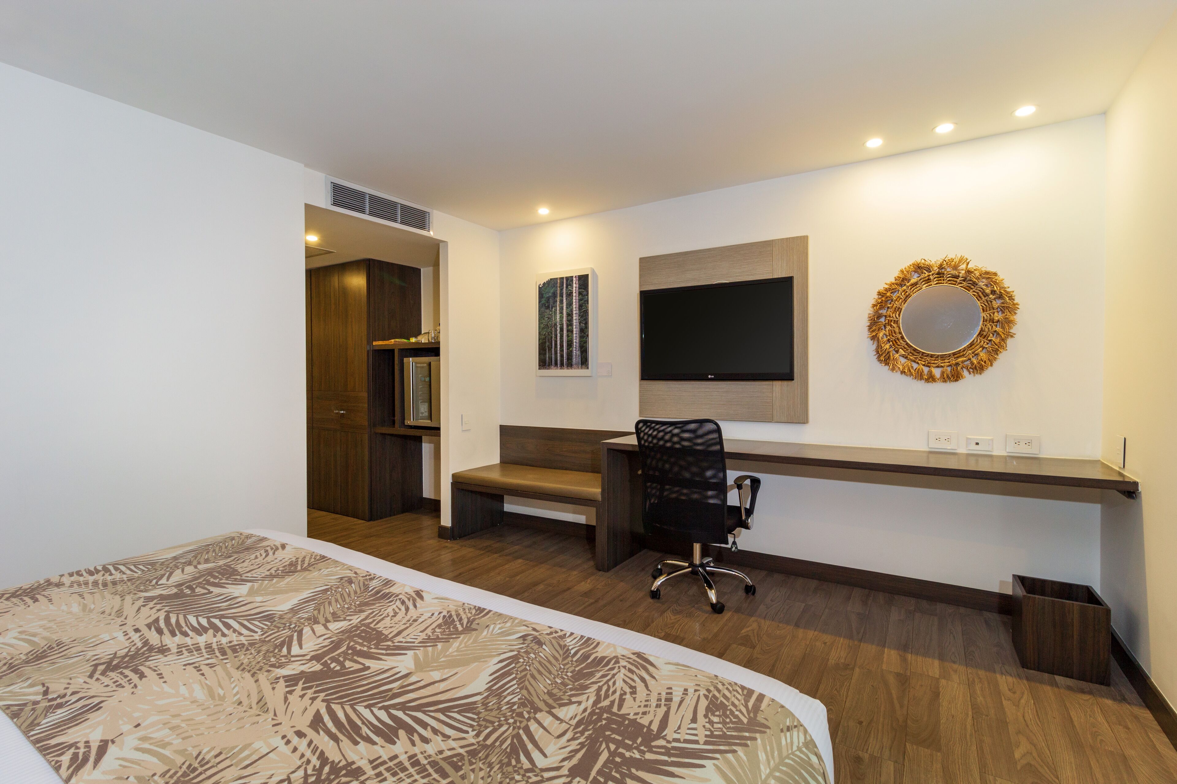 A modern hotel room with a large bed, a desk with a chair, a flat-screen TV, and a decorative mirror on the wall.