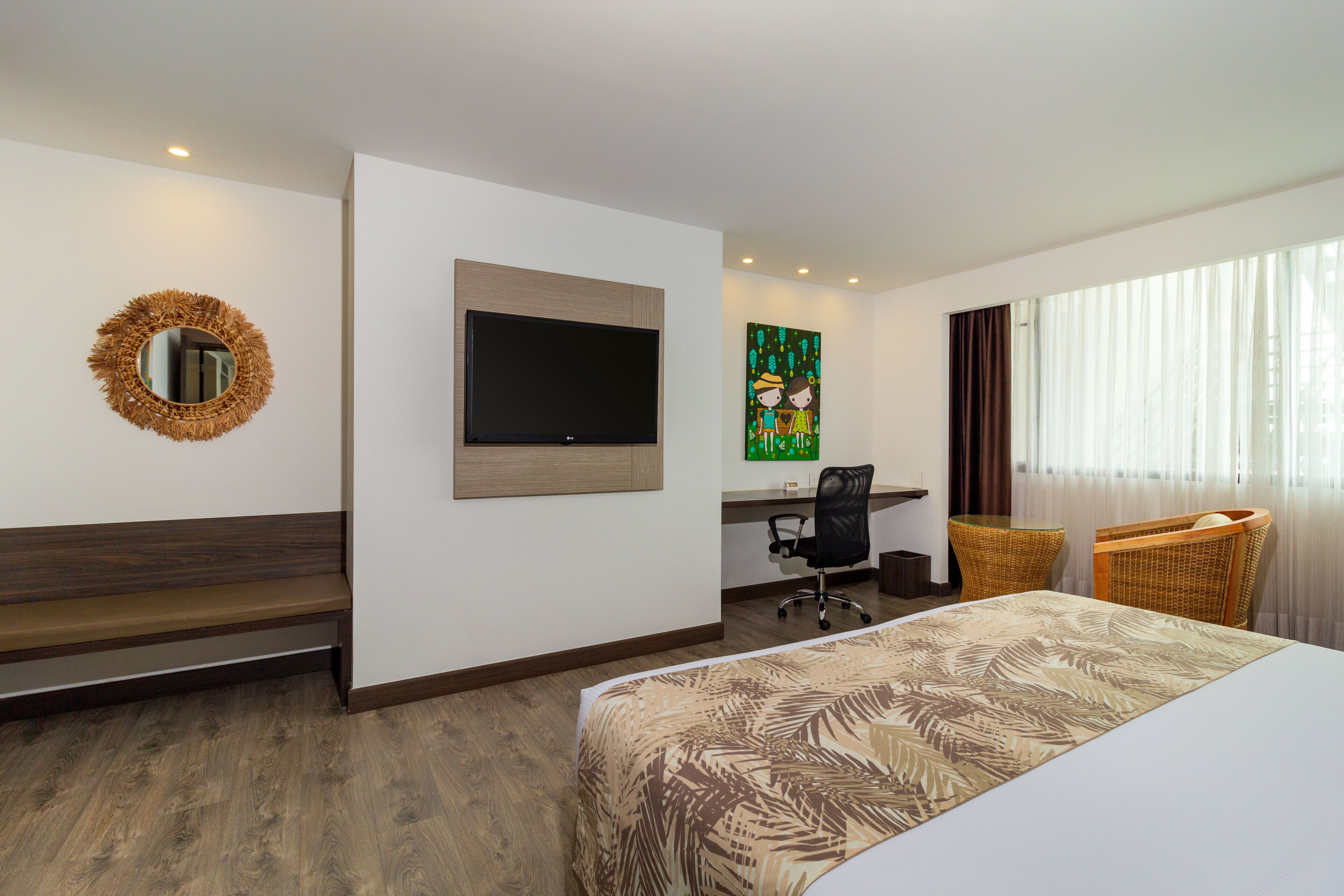 A hotel room with a bed, a desk with a chair, a television, a wicker chair, and a decorative mirror.